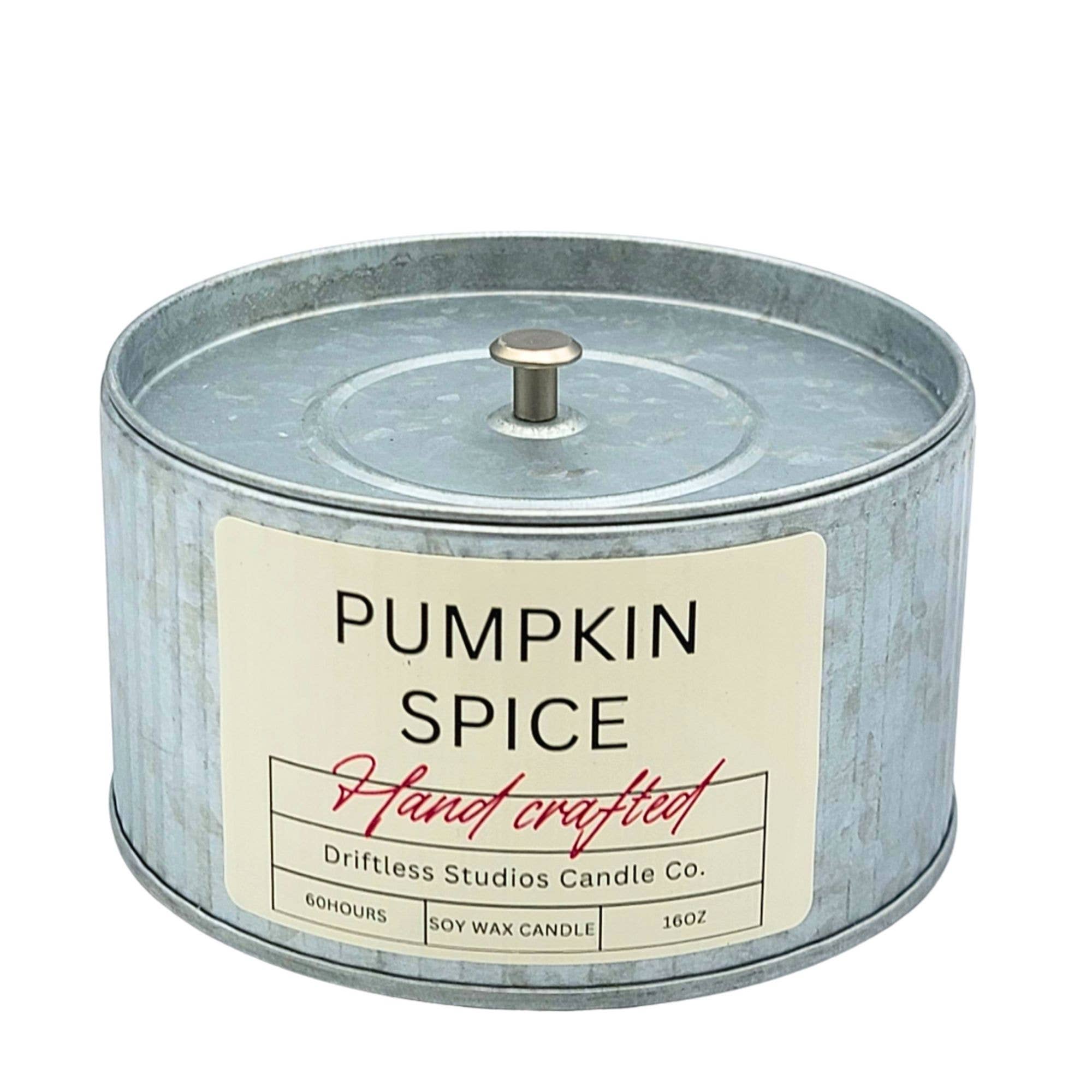 A round galvanized metal tin candle with a cream label reading 'PUMPKIN SPICE' and 'Hand crafted' in red script. The label also shows the brand 'Driftless Studios Candle Co.