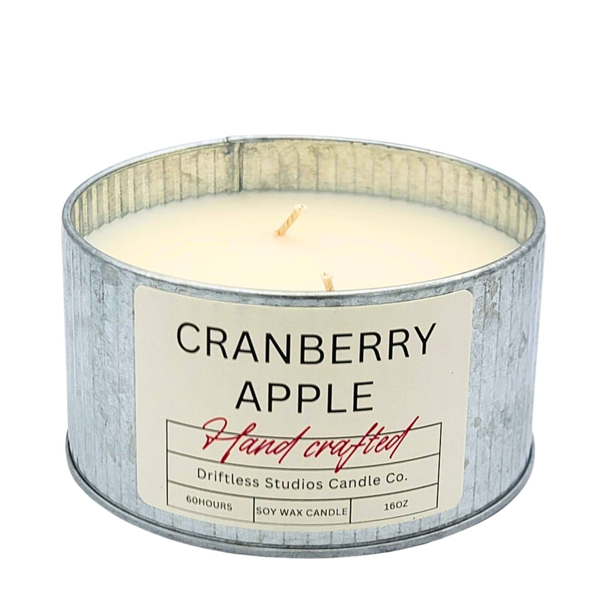A 16oz soy wax candle in a galvanized metal tin with a cream-colored label that reads 'CRANBERRY APPLE' and 'Hand crafted' by Driftless Studios Candle Co.