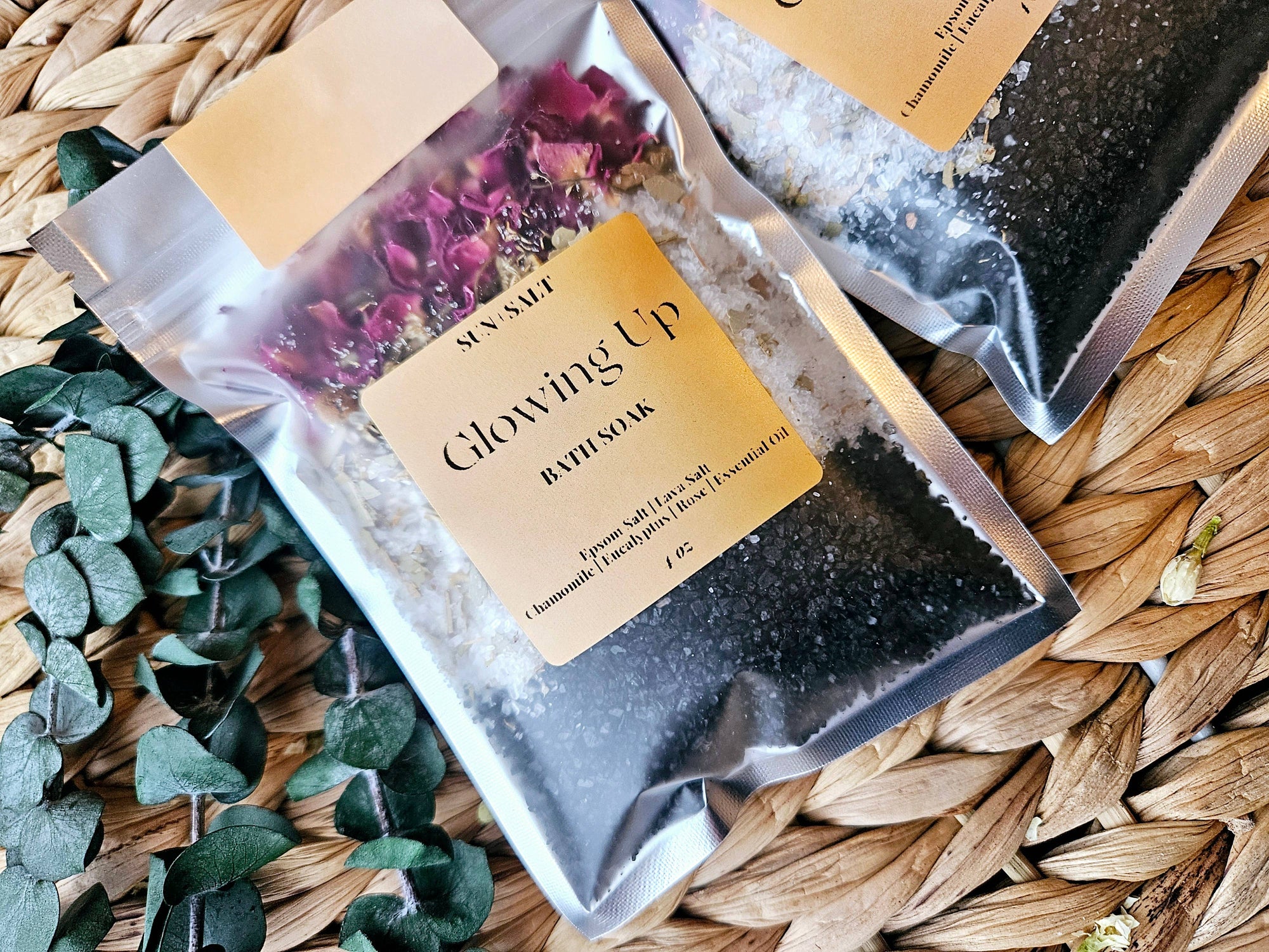 Two packages of bath soaks labeled 'Glowing Up' and 'Sun Salt' on a woven surface with greenery.