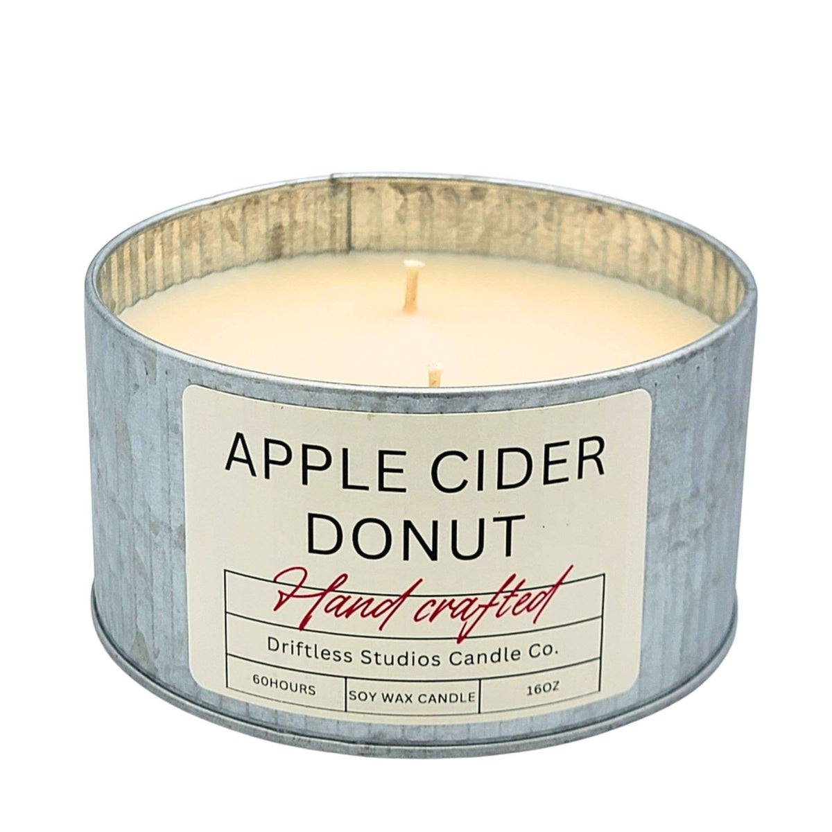 A galvanized metal tin candle with cream-colored wax, featuring a white label that reads 'APPLE CIDER DONUT' and 'Driftless Studios Candle Co.' on a plain white background.