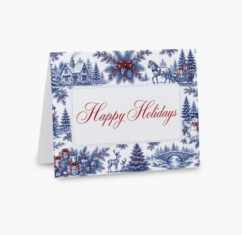 A folded holiday greeting card with a blue and red winter landscape toile de Jouy pattern, featuring the words 'Happy Holidays' in red cursive on a white center panel, shown on a light gray background.