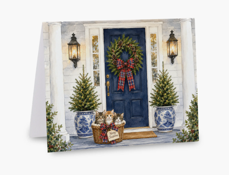 A square greeting card featuring a festive holiday scene of a blue front door with a green wreath and red plaid bow, flanked by two potted Christmas trees, with three kittens sitting in a basket on the porch. The card is shown standing upright against a plain white background.
